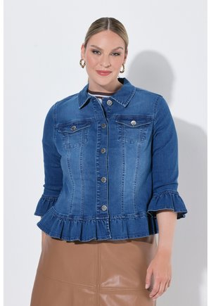 Frau mit hellem Haar trägt eine blaue Jeansjacke mit Rüschen an Ärmeln und Saum, kombiniert mit einem hellbraunen Lederrock vor einem schlichten weißen Hintergrund.