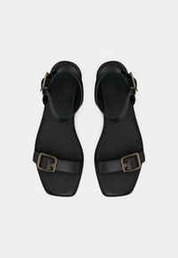 Ζευγάρι μαύρων φλατ σανδαλιών με τετράγωνη μύτη, λουριά αστραγάλου και χρυσές αγκράφες σε λευκό φόντο.