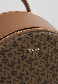 Brown leather backpack with a round shape, featuring a patterned lower section and a smooth upper section. Gold zip and branding detail.