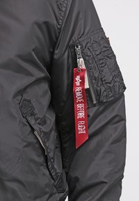 Manche de veste noire avec poche zippée et étiquette en tissu rouge indiquant "REMOVE BEFORE FLIGHT", attachée au curseur de la fermeture éclair.