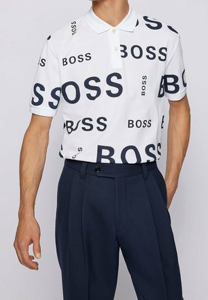 Witte polo met marineblauwe "BOSS" opdruk, korte mouwen en een puntige kraag; gecombineerd met donkerblauwe op maat gemaakte broeken met een knoopsluiting.