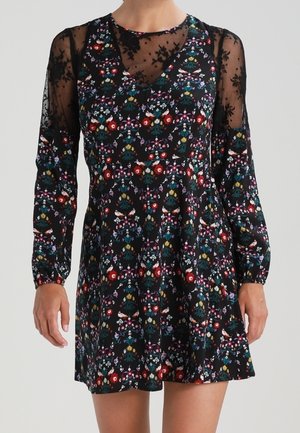 Vestito nero a fiori con maniche lunghe trasparenti e sovrapposizione trasparente sul collo, indossato da una persona in piedi visibile dalle spalle fino a metà coscia.