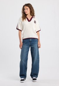 White knitted polo top featuring a deep V-neck, short sleeves with maroon accents, paired with loose blue denim jeans and black sneakers.