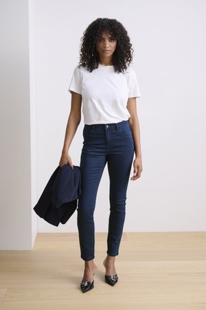 Fehér rövidujjú póló sötétkék skinny farmernadrággal párosítva. A modell egy sötétkék dzsekit tart a kezében, és hegyes fekete magassarkút visel. Az anyagok sima textúrájúak.