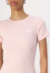 T-shirt rose clair à côtes avec des manches courtes et un col rond. Présente un petit logo brodé blanc sur le devant.