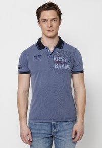 Polo majica mornarsko modre barve s ovratnikom, ki ima izvirne vezenine in grafične elemente spredaj. Kratki rokavi z kontrastnim robom in sproščen kroj.