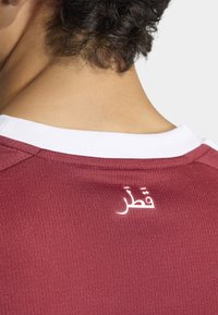 Vinröd sporttröja med vit rund halsringning; har texten "قطر" i vitt på baksidan nära kragen. Tyget har ett mönstrat struktur.