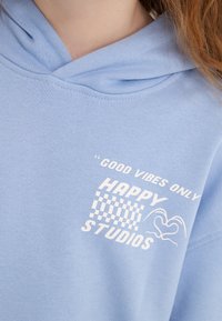 Gaiši zilais kapucis ar priekšējo kabatu, ar baltu tekstu " tikai labas vibrācijas" un "LAIMĪGIE STUDIJAS", kā arī šaha galda rakstu.