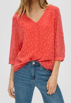 Vrouw draagt koraalrode gestructureerde blouse met driekwart mouwen en blauwe spijkerbroek, hand ontspannen langs de zijde.