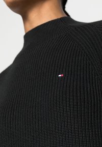 Zwart geribbelde trui met een hoge halslijn. Voorzien van een klein, meerkleurig logo op de borst. Gestructureerde stof met opvallende stiknaden.