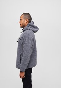 Chaqueta de forro polar gris con capucha, que presenta una superficie texturada, dos bolsillos con cremallera y un cordón ajustable en el cuello.