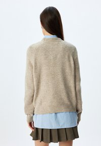 Maglione beige lavorato a maglia con scollo rotondo, polsini e bordo a coste. Indossato sopra una camicia azzurra chiara e una gonna marrone plissettata.