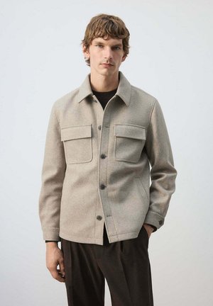 Jeune homme aux cheveux bruns courts portant une veste beige boutonnée avec des poches avant et un pantalon foncé, debout devant un fond clair uni.