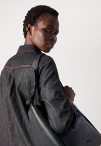 Svart lærveske med en glatt tekstur, båret over skulderen. Modellen har på seg en mørk denimskjorte med røde sømmer.