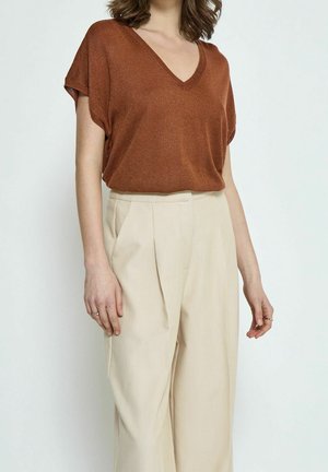 Femme portant un haut à manches courtes col en V couleur rouille, rentré dans un pantalon beige large taille haute, sur un fond uni.