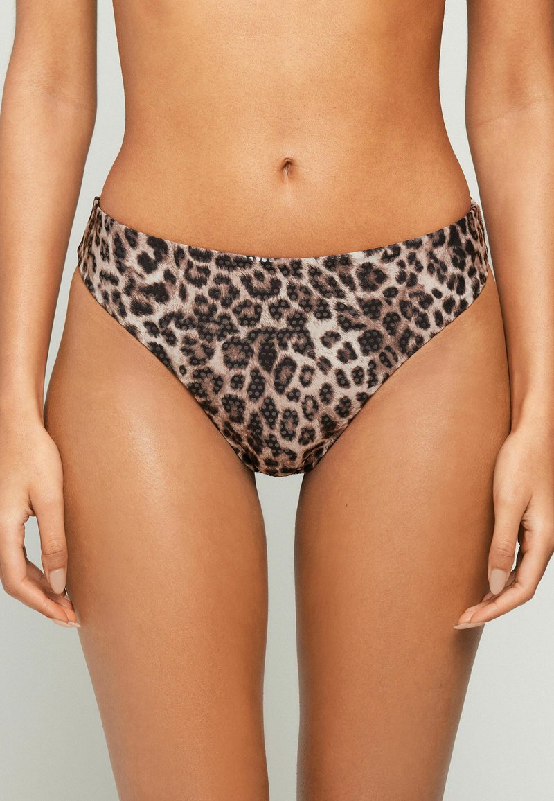 Frau trägt Leopardenmuster-Hochschnitt-Bikinihose, zeigt den Oberkörper von der Taille bis zur Mitte des Oberschenkels mit entspannten Händen an den Seiten.