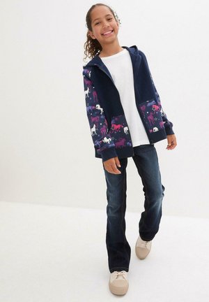 Navy Zip-Hoodie mit pinken und weißen Einhornmotiven auf den Ärmeln, kombiniert mit dunklen Jeans und beigen Sneakers, stehend auf einem weißen Boden.