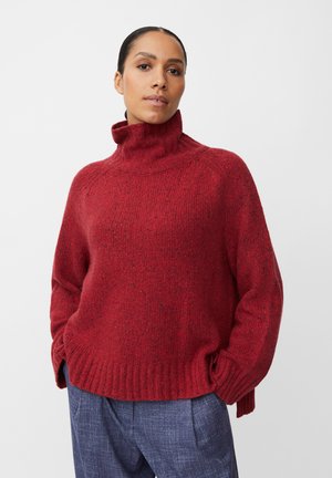 Roter Strickpullover mit hohem Kragen, strukturiertem Muster und gerippten Bündchen. Kombiniert mit lockeren blauen Hosen.