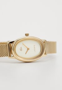 Montre-bracelet en mailles dorées avec un cadran ovale, un cadran ivoire, une lunette dorée texturée et le logo Lacoste. Aiguilles simples pour les heures et les minutes.
