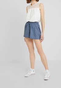 Top bianco senza maniche con dettagli in pizzo, abbinato a pantaloni corti blu con motivo e scarpe da ginnastica bianche alte, con vita a cordino.