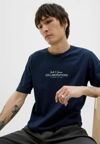 T-paita tummansinistä puuvillaa, jossa on valkoinen "Jack & Jones COLLABORATIONS" -grafiikka, pyöreä kaula-aukko, lyhyet hihat, yhdistetty tummiin housuihin.