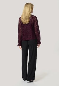 Top in pizzo a maniche lunghe di un profondo bordeaux, con motivi floreali e una vestibilità ampia, abbinato a pantaloni neri a gamba larga e tacchi neri.