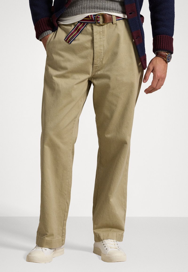 Pantalons en coton beige avec une coupe décontractée, dotés d'une ceinture rayée et associés à des chaussures blanches, affichant un look décontracté.