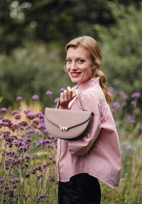 Růžová kožená kabelka zakřiveného tvaru se zlatými kovovými prvky a ramenním popruhem, kterou drží osoba stojící mezi květinami.