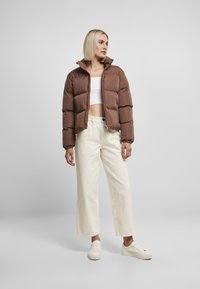 Brun puffet jakke med høj krave, lynlås foran og elastiske manchetter, parret med cremede brede bukser og hvide sneakers.