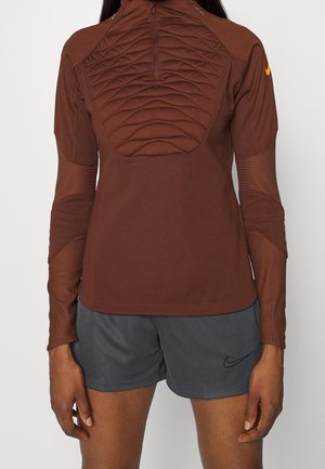 Personne portant un haut de sport marron texturé à manches longues avec une fermeture éclair au quart et un short Nike gris avec un logo Swoosh noir.