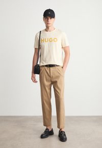 T-shirt beige avec "HUGO" en orange vif, pantalon beige, et mocassins noirs. Les accessoires incluent une casquette noire et un petit sac bandoulière.