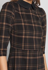 Svart och brun rutig klänning med krage, trekvartslånga ärmar och en slät textur. Har rutiga mönsterdetaljer.