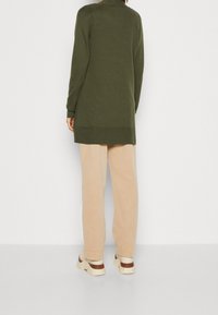 Maglione verde lungo con orlo a coste, abbinato a pantaloni beige e scarpe chiare. Texture morbida e vestibilità rilassata.