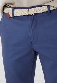 Navyblaue Hose mit einem gewebten cremefarbenen Gürtel. Der Gürtel hat ein braunes Lederakzent. Elegante, glatte Textur und taillierte Passform.