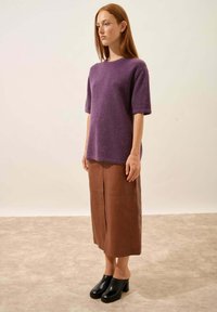Fioletowa dzianinowa bluzka z krótkim rękawem połączona z brązową skórzaną spódnicą midi i czarnymi mule na obcasie, na jednolitym tle.