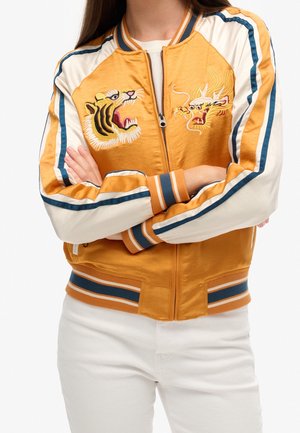 Mujer que lleva una chaqueta bomber en color mostaza y crema con bordados de tigre y dragón, rayas azules y blancas en las mangas, y pantalones blancos.