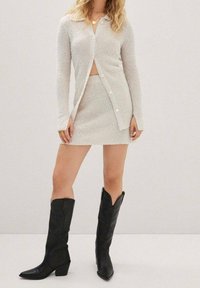 Gilet en tricot gris clair avec des boutons, associé à une mini jupe en tricot assortie. Bottes de cowboy en cuir noir, montantes jusqu'aux genoux, avec des bouts pointus.