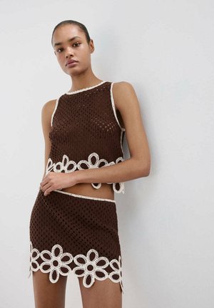 Joven mujer que lleva un top marrón de ganchillo sin mangas y una minifalda a juego con adornos florales blancos, de pie contra una pared blanca.