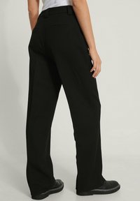 Black wide-leg trousers made of textured fabric. Two back pockets and a tailored waistband with belt loops. Pants are paired with black shoes.