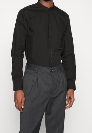 Mann trägt ein schwarzes Langarmshirt mit verdeckter Knopfleiste und graue plissierte Hose vor einfarbigem Hintergrund.