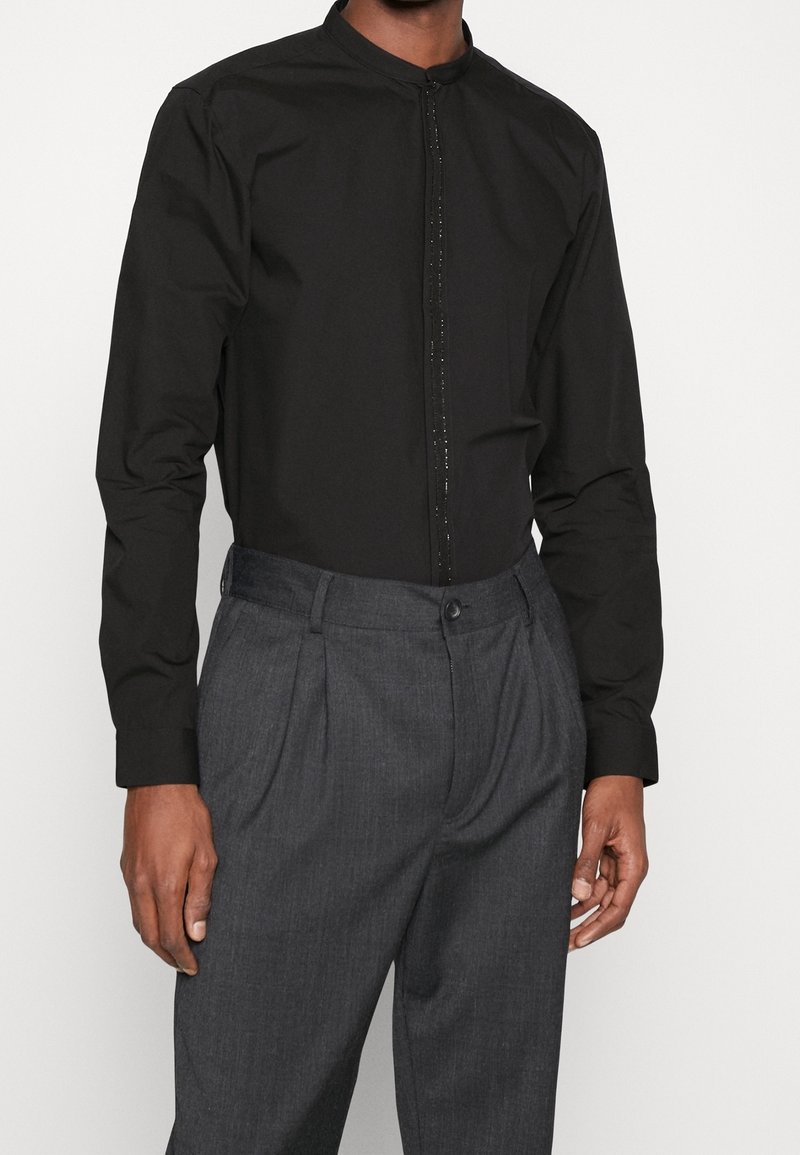 Homme portant une chemise noire à manches longues avec patte de boutonnage cachée et un pantalon plissé gris sur un fond uni.