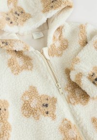 Weiße Fleece-Jacke mit Reißverschluss und Kapuze, die mit beigen Teddy-Muster verziert ist. Sie hat eine weiche Textur und einen cremefarbenen Reißverschluss. Mit dem Label "NEXT Baby."