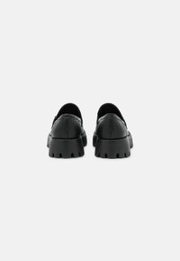 Zapatos slip-on de cuero negro con suela de goma gruesa que presenta cortes profundos y un acabado suave, vista trasera que muestra un diseño minimalista.