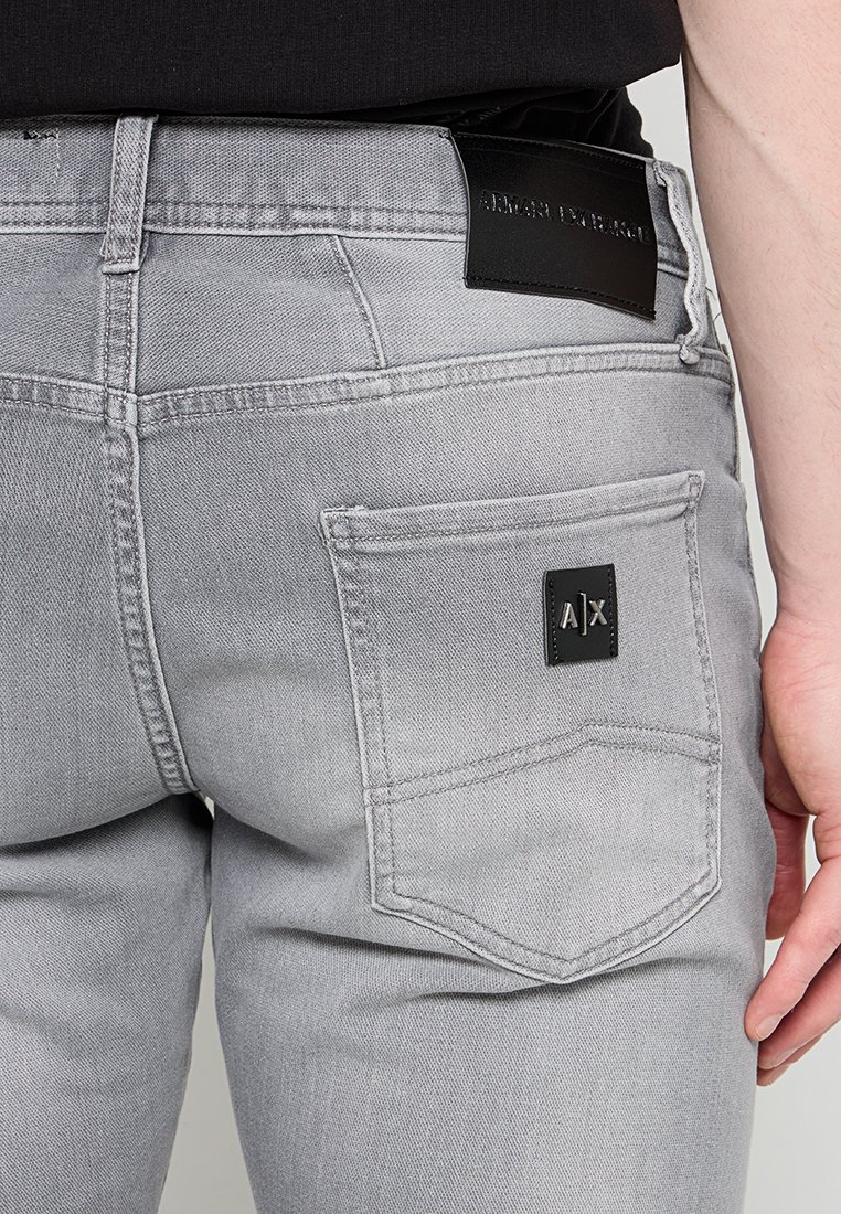 Szare dżinsy mają tylną kieszeń z czarną łatą z logo. Wygląd obejmuje standardowe szwy i klasyczny krój.