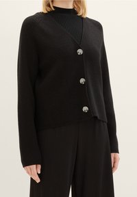 Černý žebrovaný cardigan s výstřihem do V, dlouhými rukávy a čtyřmi stříbrnými knoflíky vpředu. Měkká textura, volný střih.