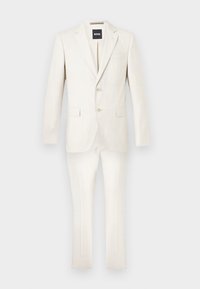 Κρεμ κοστούμι δύο τεμαχίων με σακάκι δύο κουμπιών και ταιριαστό παντελόνι στενής εφαρμογής σε απλό φόντο.