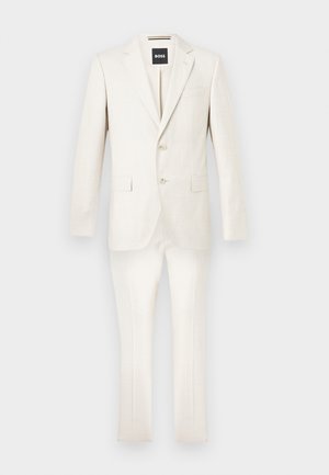 Dwuczęściowy garnitur w kremowym kolorze z marynarką na dwa guziki i dopasowanymi spodniami wąskiego kroju na jednolitym tle.