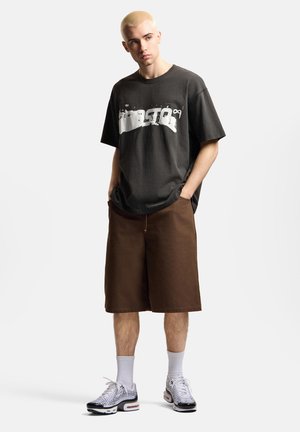 Młody mężczyzna z krótkimi blond włosami, ubrany w czarną koszulkę z nadrukiem, brązowe szorty za kolana, białe skarpetki oraz czarno-białe trampki.