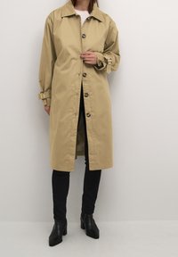 Beżowy płaszcz trench z guzikiem z przodu, szerokim kołnierzem i mankietami. Wykonany z gładkiego materiału, z długością do kolan i bocznymi rozcięciami.
