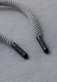 Gros plan sur des embouts de lacets tressés gris avec des aglets en plastique noir gravés de "OMORE" sur un fond en tissu gris texturé.
